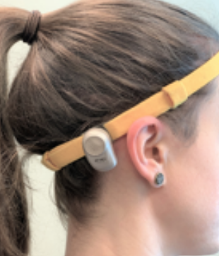 A person with brown hair in a ponytail wears a yellow headband with a small gray electronic device attached above their ear.