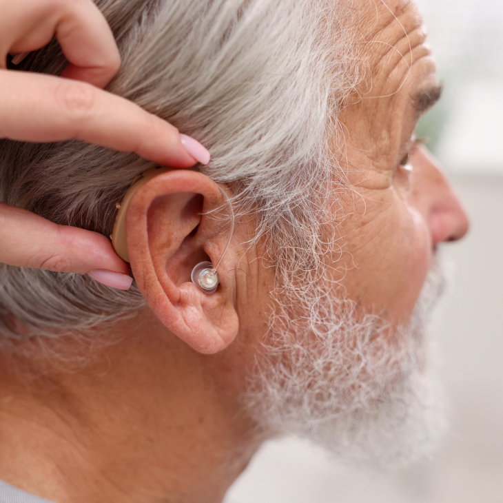 A person with gray hair and a beard wears a hearing aid behind their ear, highlighting the importance of hearing aids in managing hearing loss, while another hand helps adjust it.