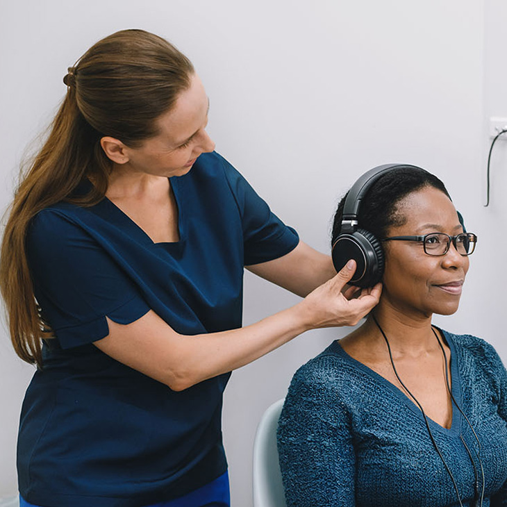 A healthcare professional fits large headphones on a seated woman with glasses, likely preparing her for a hearing test in a clinical setting—an important step in evaluating hearing loss and determining the need for hearing aids.