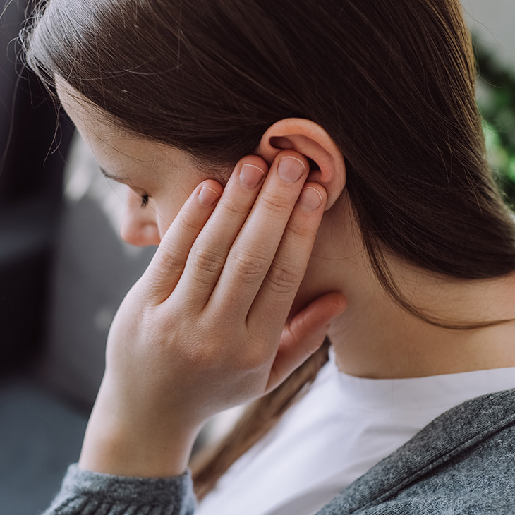 A person with long brown hair touches their ear with one hand, appearing to experience ear discomfort or pain, possibly indicating hearing loss or the need for hearing aids.